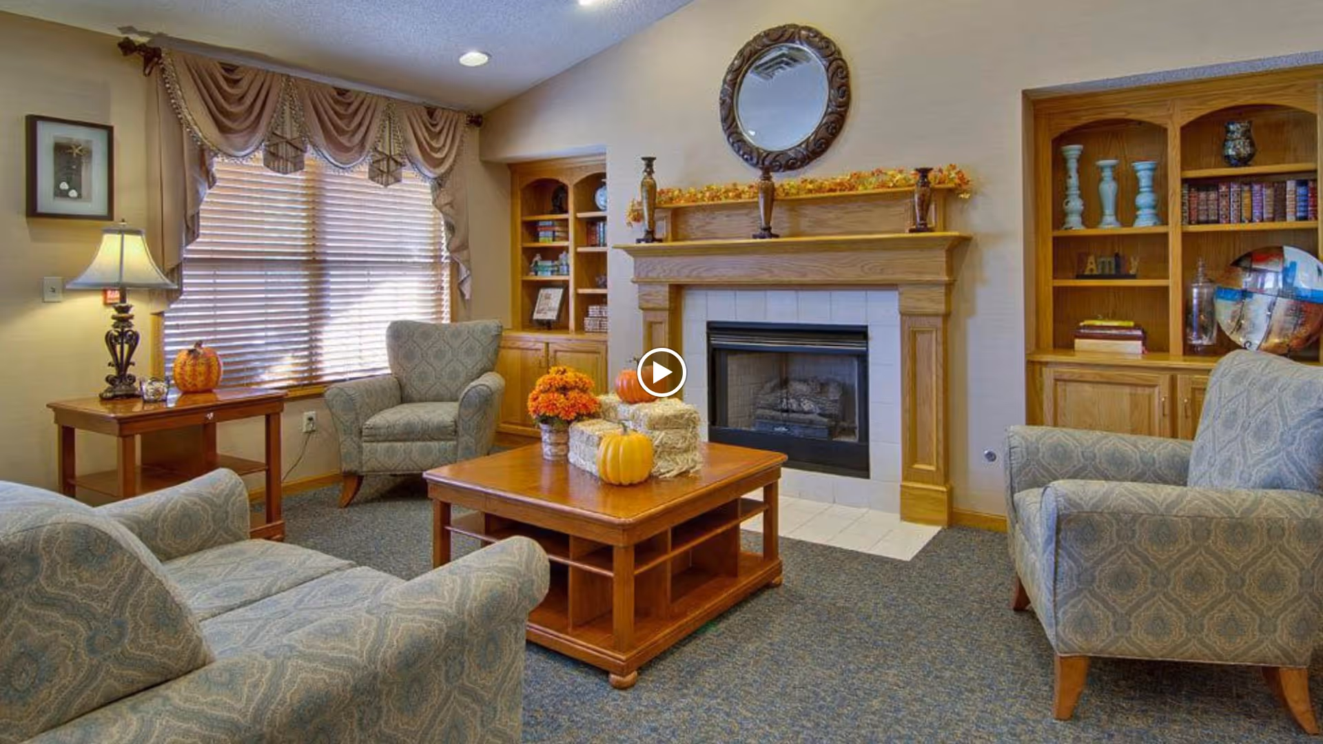 A cozy senior living room with three upholstered armchairs arranged around a wooden coffee table decorated with small pumpkins and flowers. The room features a fireplace with a wooden mantel adorned with autumn decorations and a round mirror above it. Built-in wooden shelves flank the fireplace, holding books and decorative items. A window with blinds and draped curtains lets in natural light, and a table lamp sits on a side table next to one of the chairs.