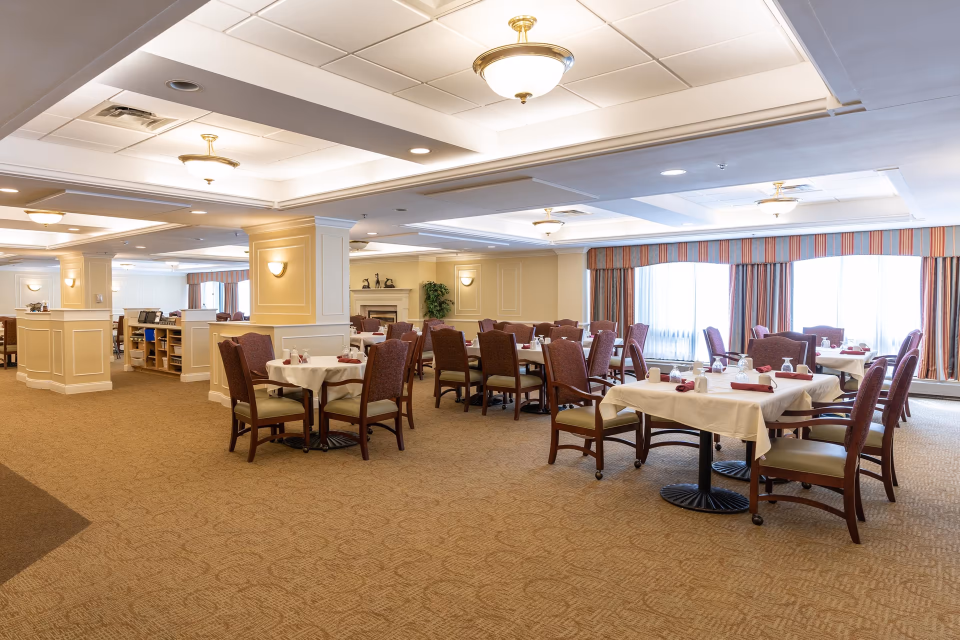 A spacious dining room with multiple tables covered in white tablecloths, each set with napkins and glassware. The room features carpeted floors, upholstered chairs, large windows with striped curtains, and soft ceiling lighting. The walls are painted in a light beige color with decorative molding and sconces, creating a warm and inviting atmosphere.
