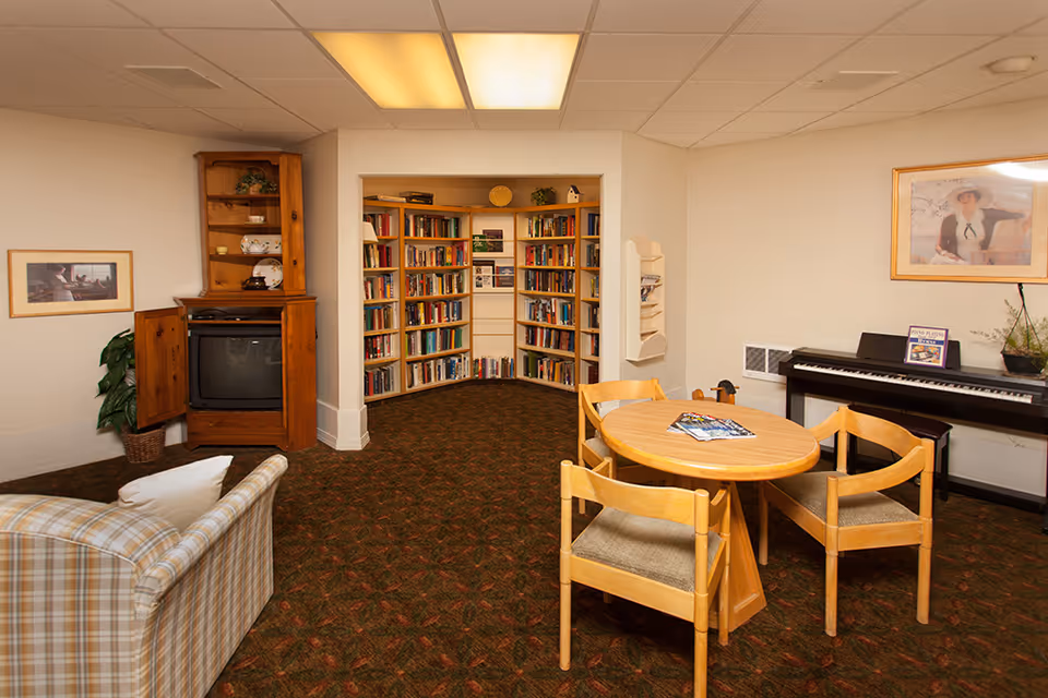 A cozy common area in a senior living facility featuring a round wooden table with four chairs, a piano against the wall with sheet music on it, a built-in bookshelf filled with books, a wooden cabinet with a TV inside, a plaid armchair, framed artwork on the walls, and a patterned carpet.