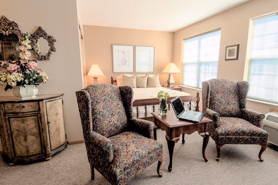 A cozy senior living bedroom with a large bed in the background, two floral upholstered armchairs facing a small wooden table with a laptop and a vase of flowers on it. The room has beige walls, two framed pictures above the bed, two table lamps on either side of the bed, a decorative cabinet with a floral arrangement, and two large windows letting in natural light.