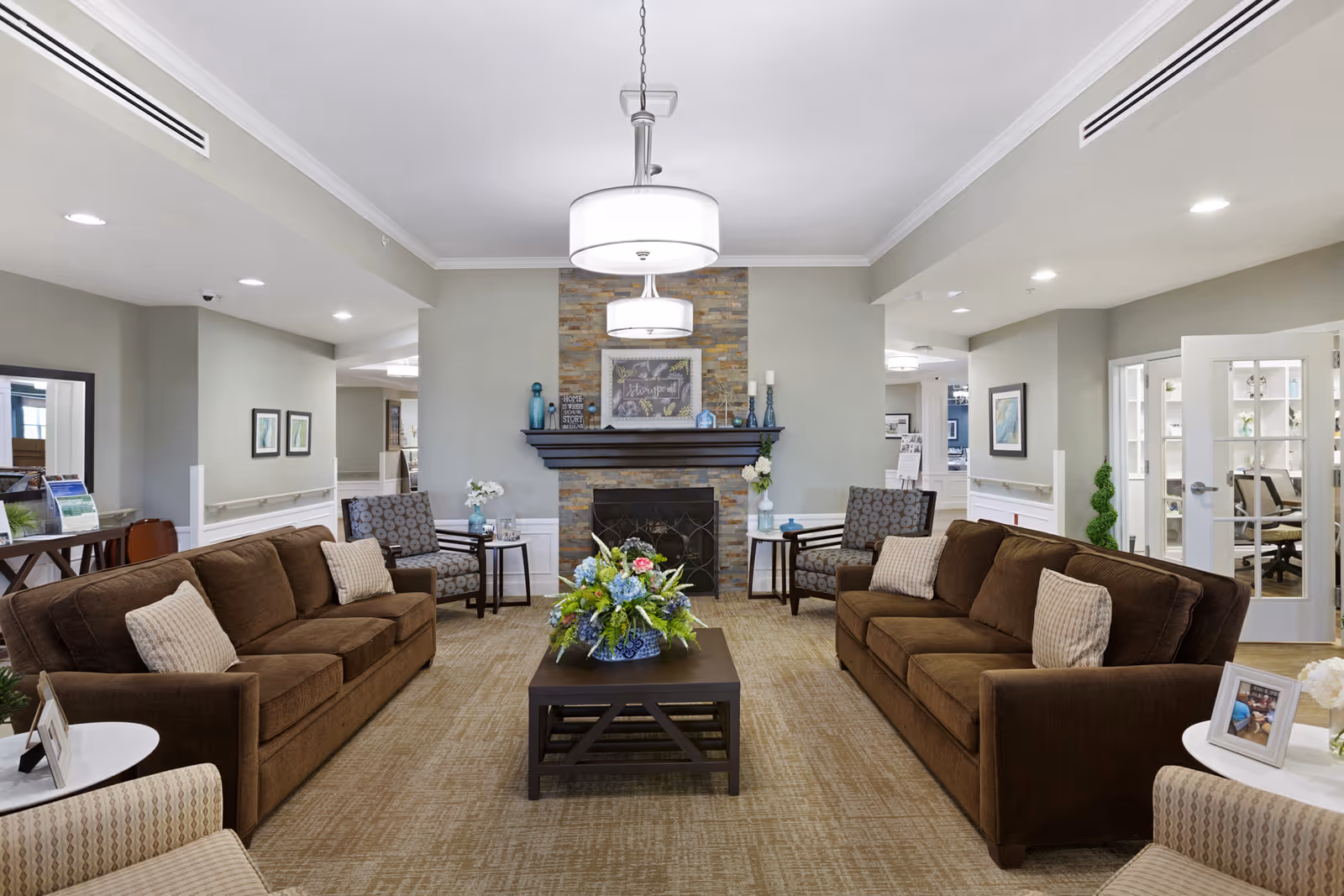 A cozy living room area in a senior living facility with two brown sofas facing each other, a dark wooden coffee table with a floral arrangement in the center, two patterned armchairs with side tables, and a stone fireplace with decorative items on the mantel. The room has beige carpet, soft lighting from ceiling fixtures, and light gray walls with white trim.