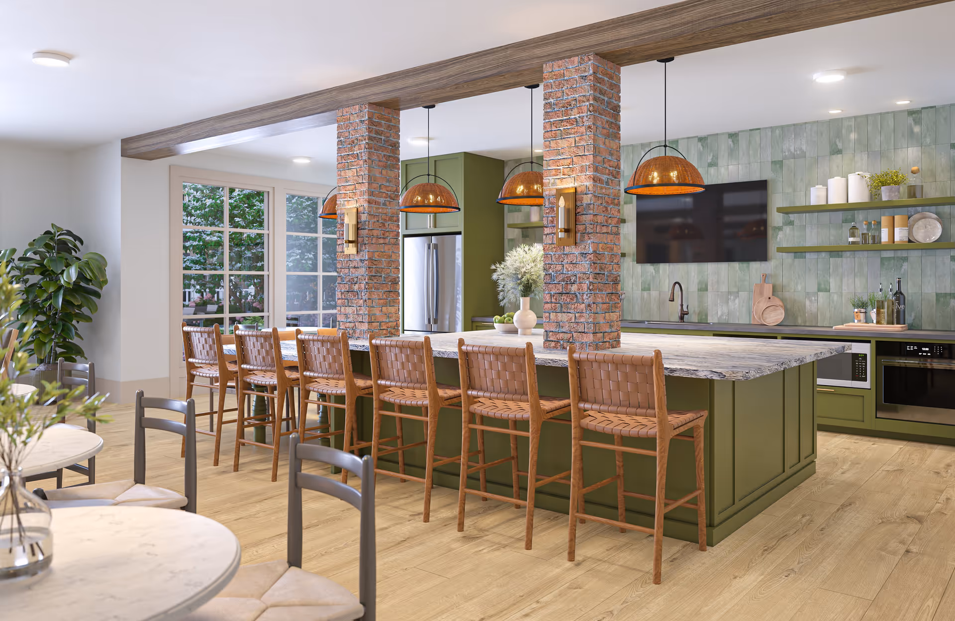 Modern kitchen and dining area with a large green island featuring a marble countertop and seven woven leather bar stools. The kitchen has green cabinetry, a stainless steel refrigerator, built-in microwave, and oven. Three pendant lights with amber shades hang above the island. There are two brick columns supporting a wooden beam above the island. In the foreground, there are round marble tables with gray chairs. A large window shows greenery outside, and a flat-screen TV is mounted on a green tiled wall with open shelves holding decorative items.