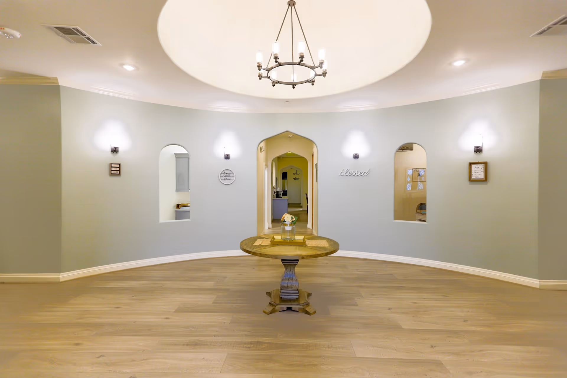 Bright circular lobby with a central round table beneath a chandelier, arched doorway and curved wall with small windows and sconces.