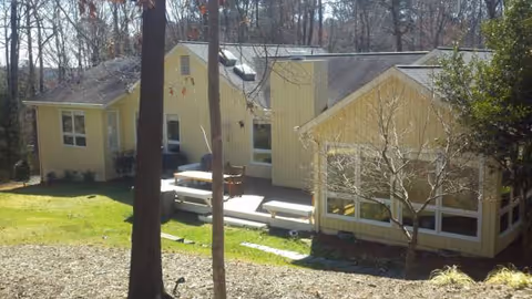 Yellow, single-story senior living building with outdoor patio benches and surrounding trees.