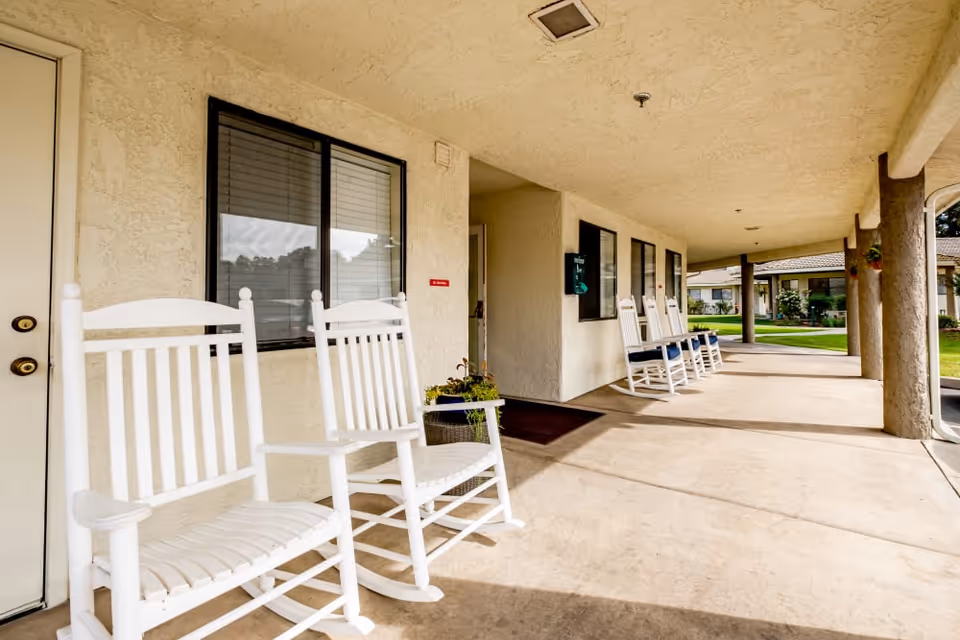 Covered exterior walkway with white rocking chairs along the entrance of a senior living facility.