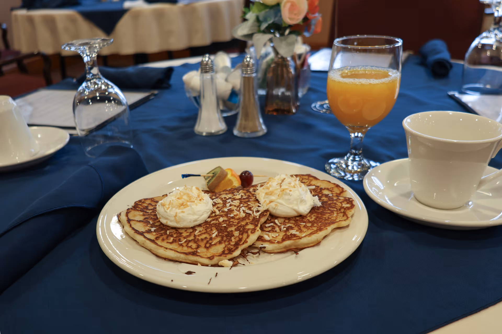 A plate with two pancakes topped with whipped cream and toasted coconut flakes, garnished with a slice of kiwi, orange, and a cherry, placed on a table with a blue tablecloth. Next to the plate is a glass of orange juice and a white cup and saucer. In the background, there are salt and pepper shakers, a small flower arrangement, and an upside-down wine glass.