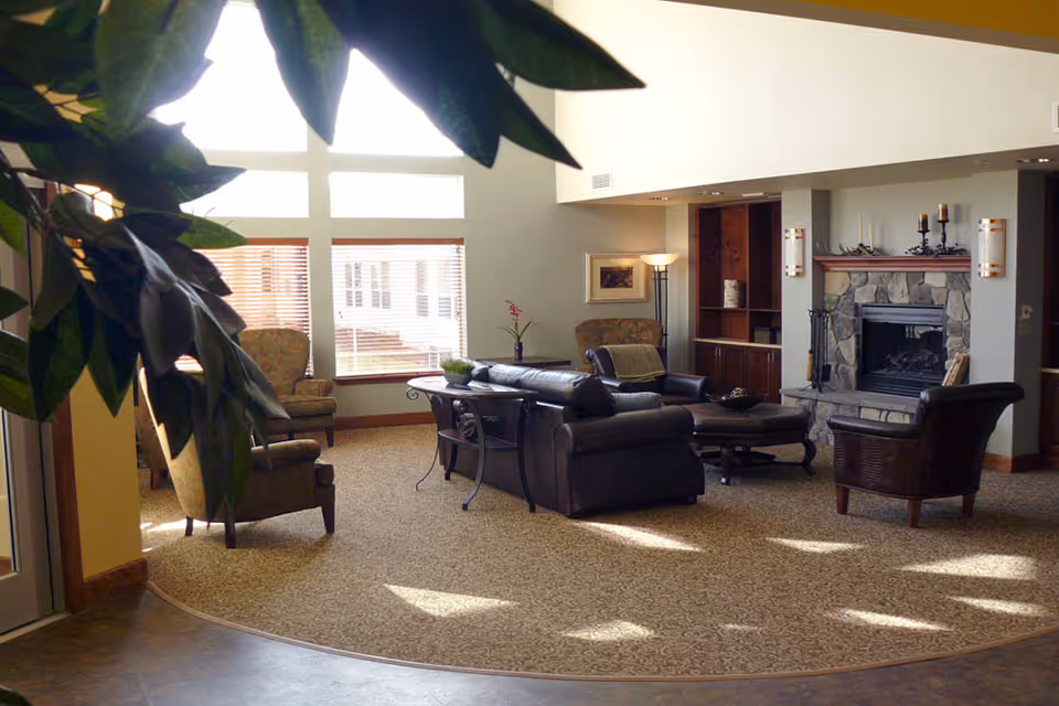 A cozy living room area with a large window letting in natural light. The room features a dark leather sofa, two armchairs, a stone fireplace, a floor lamp, and a small table with a plant. There are built-in wooden shelves and a carpeted floor with a patterned rug. Some green leaves from a plant are visible in the foreground.