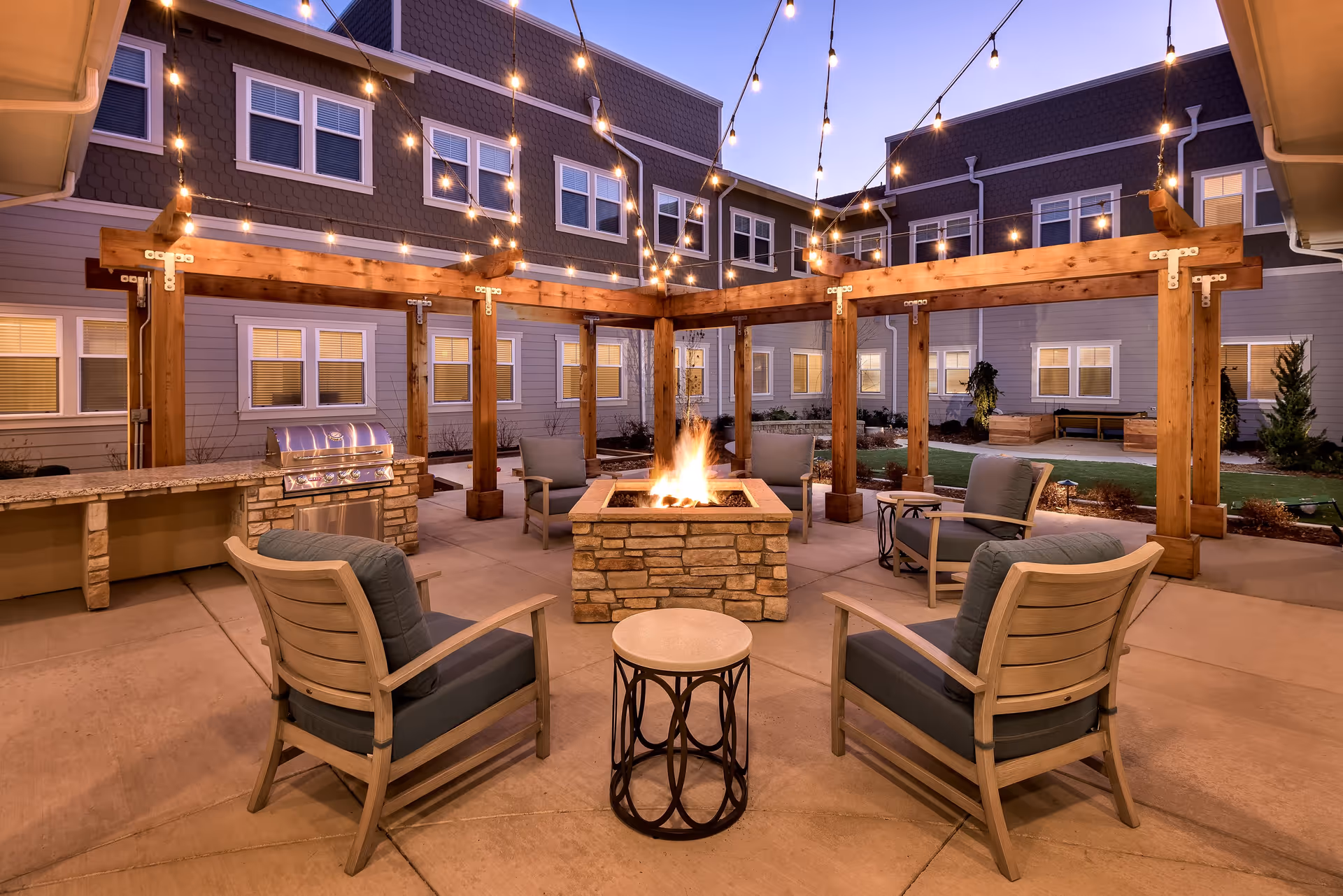 Outdoor courtyard at dusk featuring a central stone fire pit surrounded by cushioned chairs, a wooden pergola with string lights, and a built-in grill.