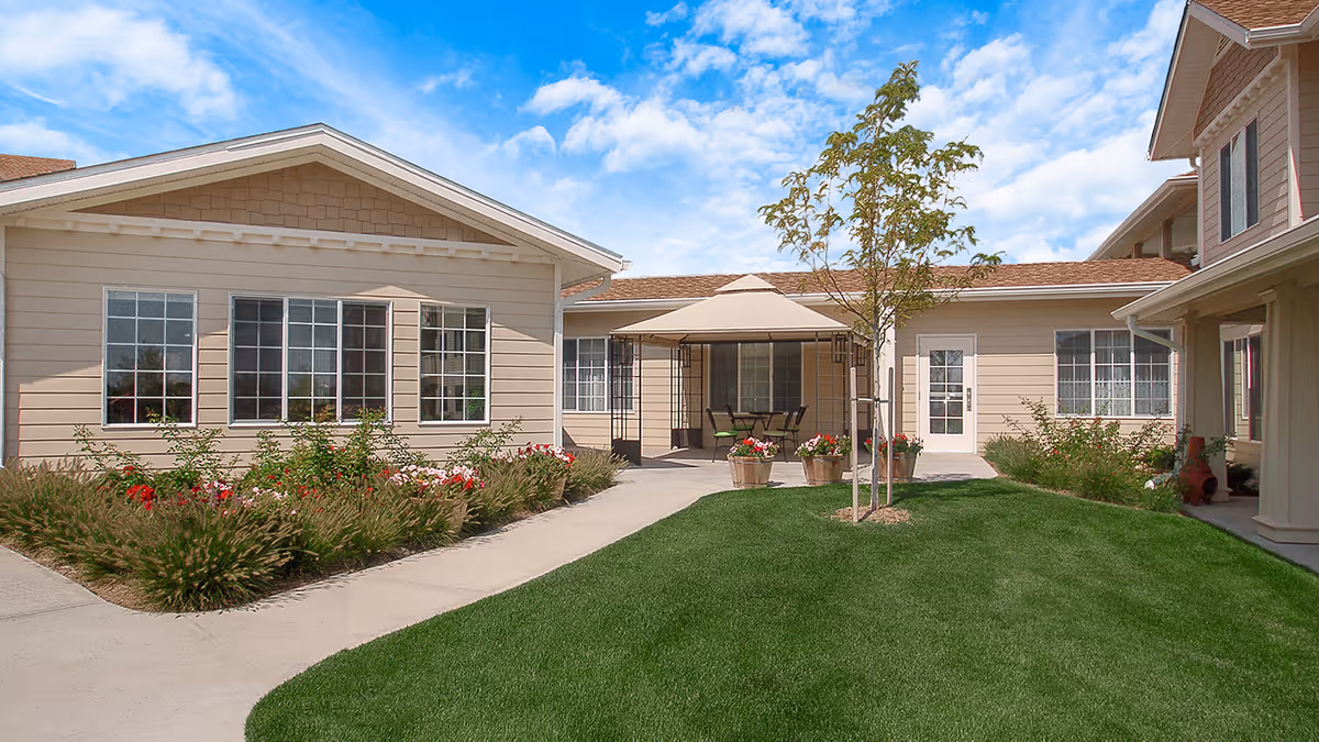 Courtyard of a beige senior living facility with a green lawn, paved walkways, flower beds, a small tree and a shaded patio with chairs.