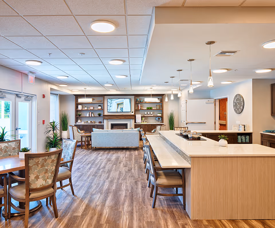 A spacious and well-lit common area in a senior living facility featuring a large kitchen island with seating, a dining table with chairs, a cozy seating area with a sofa and armchairs, a fireplace, and built-in shelves with decorative items. The room has wood flooring, pendant lights, and large windows allowing natural light.