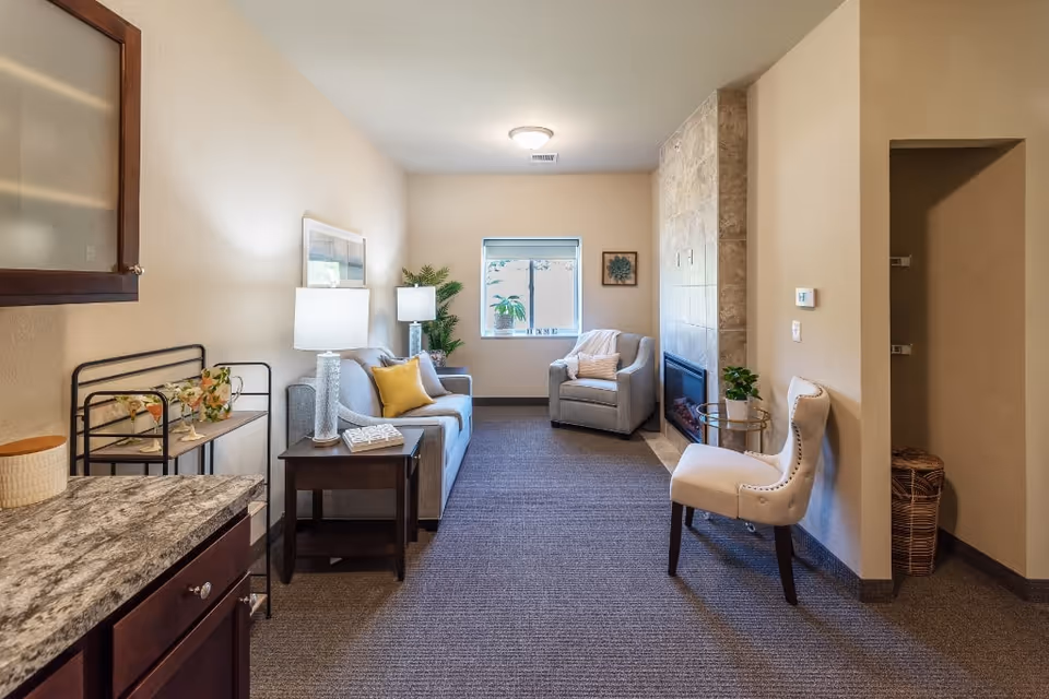 Furnished living room with a sofa, armchair, side tables, lamp, and a fireplace beneath a tiled accent wall.
