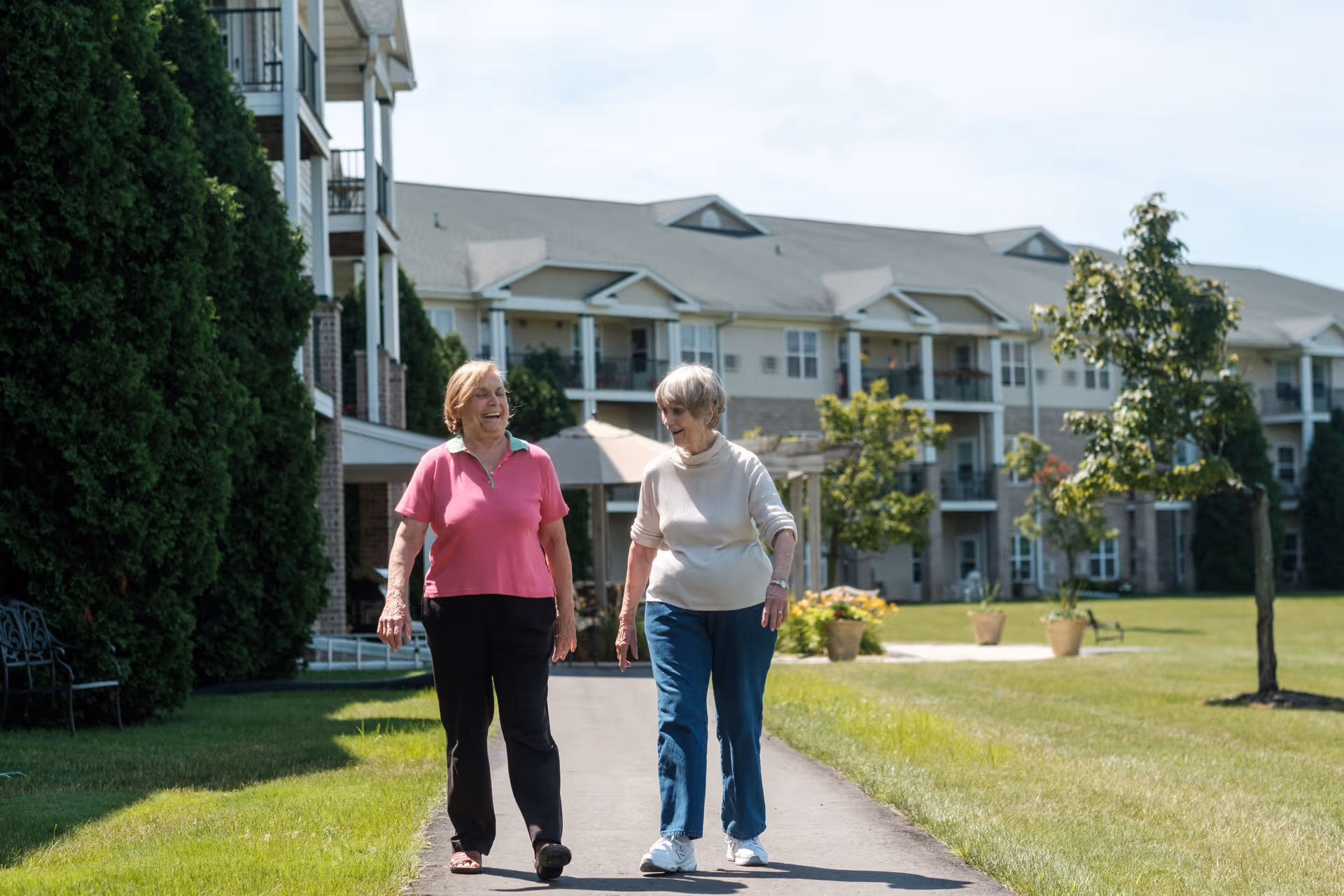 Two elderly women walking and talking on a paved path outside a multi-story assisted living building with balconies, surrounded by green grass, trees, and potted plants on a sunny day.