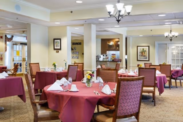 A dining room in a senior living facility with multiple tables covered in burgundy tablecloths, each set with white napkins, cups, and small flower arrangements. The room has beige walls, framed artwork, and chandeliers hanging from the ceiling. There is a counter area in the background with some items on it.