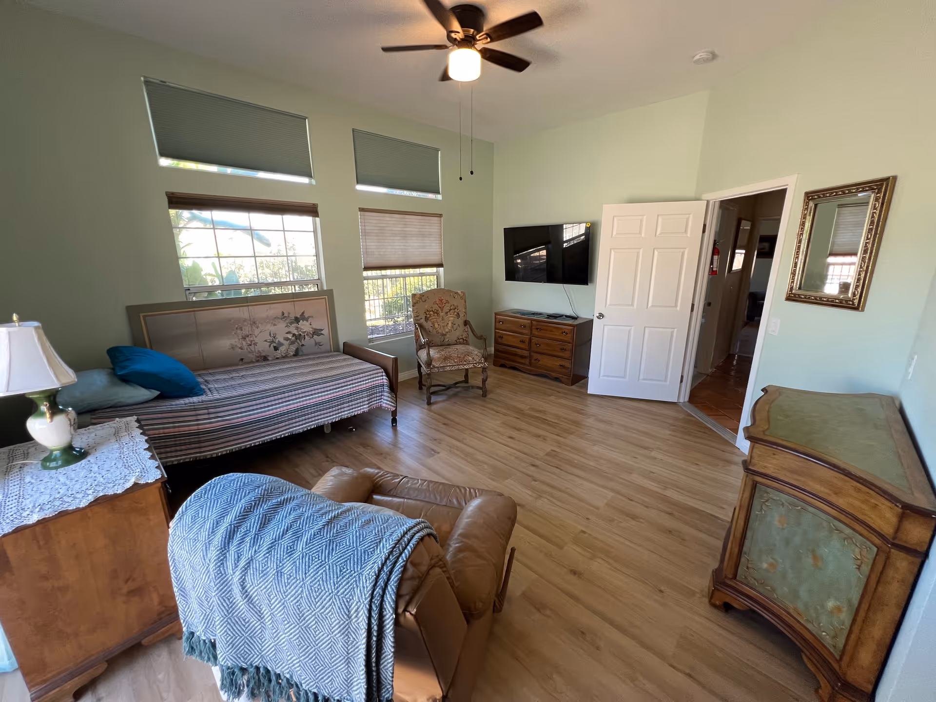 Bright furnished bedroom with a daybed, recliner, armchair, wall-mounted TV, wooden dressers and a ceiling fan.
