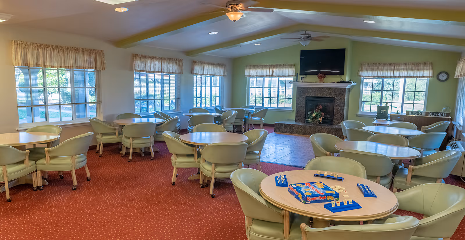 A spacious common room with multiple round tables surrounded by green cushioned chairs on a red carpet. Large windows with beige curtains let in natural light. A fireplace with a TV mounted above it is visible at the far end of the room. One table in the foreground has a Rummikub game set up. A sign reading 'Bingo Special' is on a table near the windows.