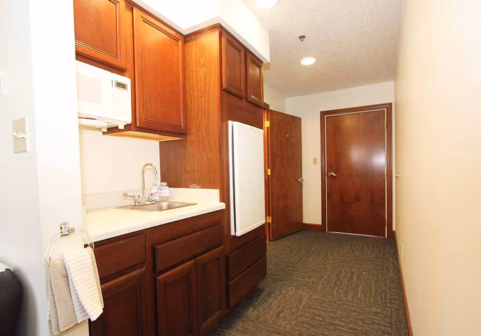 Small kitchenette with wooden cabinets, a sink, microwave, and compact refrigerator along a carpeted hallway leading to two wooden doors.