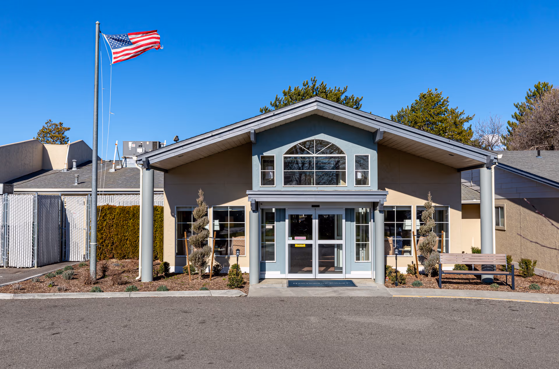 Front exterior view of Hawthorne Court facility building with a peaked roof, large windows, a bench on the right side, and an American flag on a flagpole to the left. The sky is clear and blue.