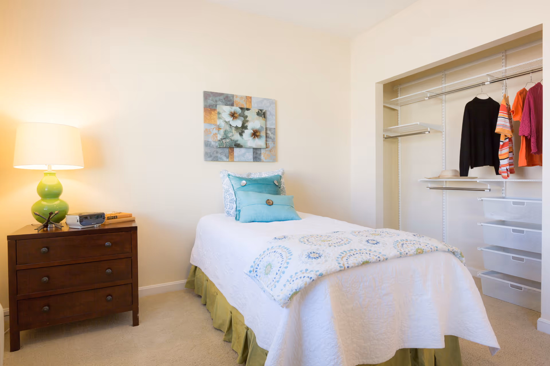 A small bedroom with a single bed covered in a white quilt and decorative pillows in shades of blue and green. To the left of the bed is a dark wooden nightstand with a green lamp, a clock, and some books. On the wall above the bed is a floral artwork. To the right is an open closet with hanging clothes and white storage drawers.