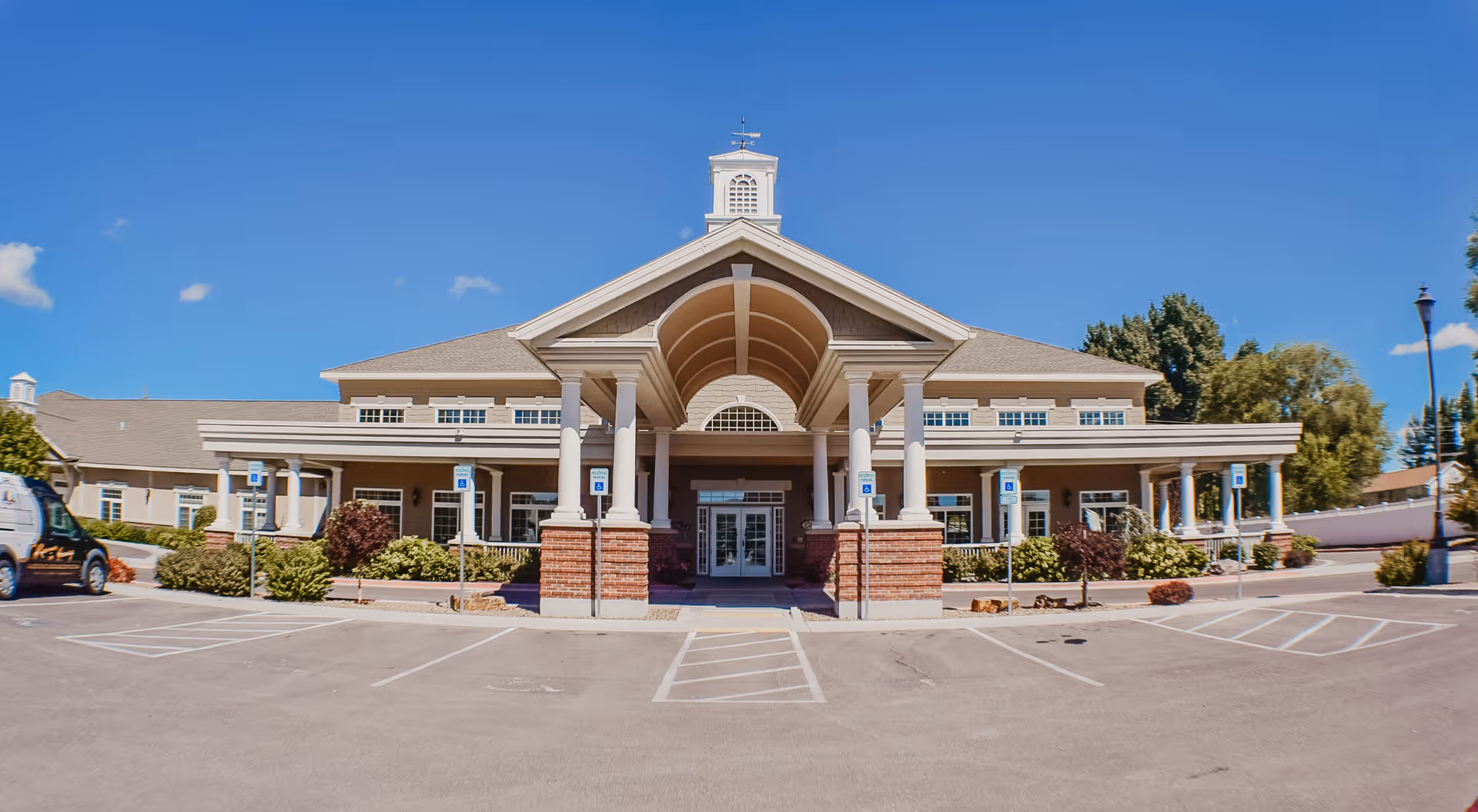 Front exterior view of Carriage Cove Short Stay Rehabilitation building with a covered entrance supported by white columns, brick accents, and a clear blue sky above. Handicap parking spaces are visible in front of the building along with some landscaping and a parked van on the left side.