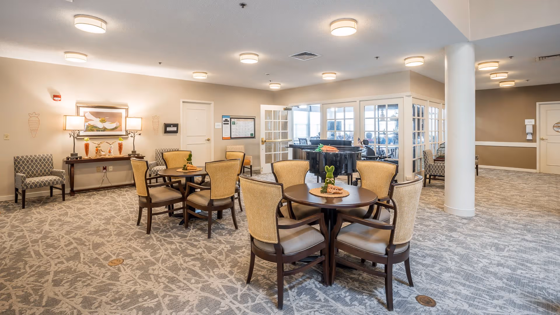 A spacious, well-lit common area with round tables and cushioned chairs arranged for seating. The room features a patterned carpet, soft beige walls, and ceiling lights. There is a console table with lamps and decorative items against one wall, and a black grand piano is visible in an adjacent room through glass-paneled doors.