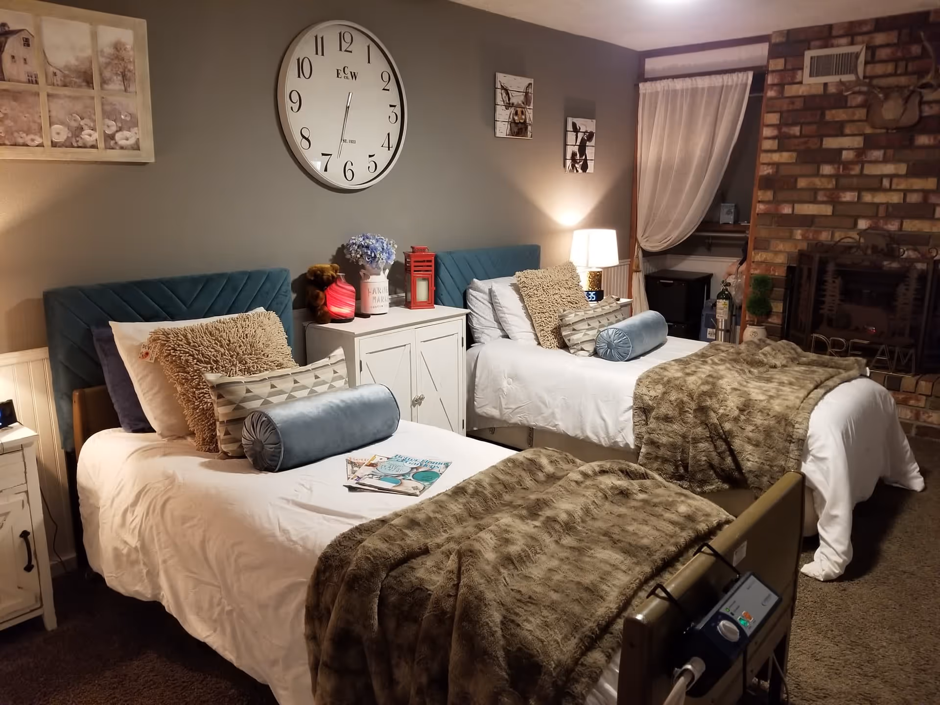 A cozy bedroom with two single beds, each with white bedding, decorative pillows, and brown fur throws. Between the beds is a white nightstand with a teddy bear, a red lantern, and a vase with blue flowers. Above the nightstand is a large wall clock. The room has a brick fireplace on the right side and soft lighting from a table lamp on the nightstand. The walls are decorated with framed pictures and the room has a warm, inviting atmosphere.