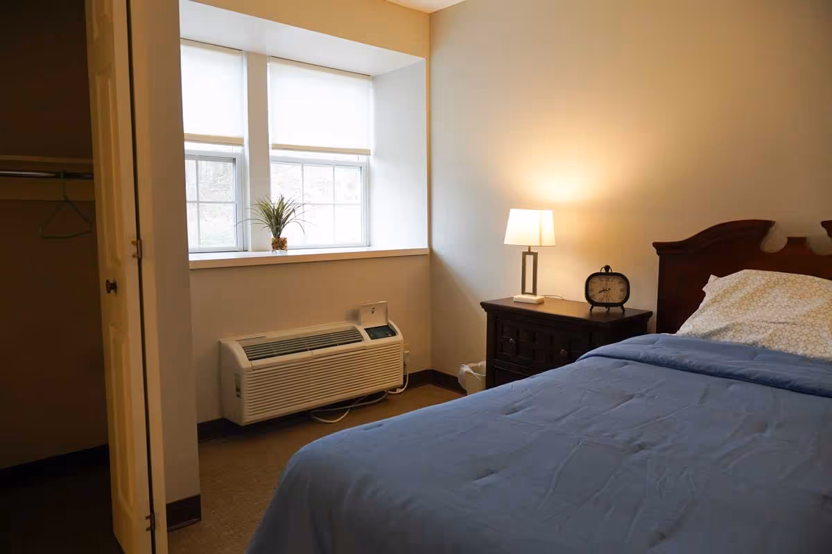 A cozy bedroom with a bed covered in a blue blanket and white pillow, a wooden nightstand with a lamp and an alarm clock, a window with white blinds, a small plant on the windowsill, and an open closet with hangers.