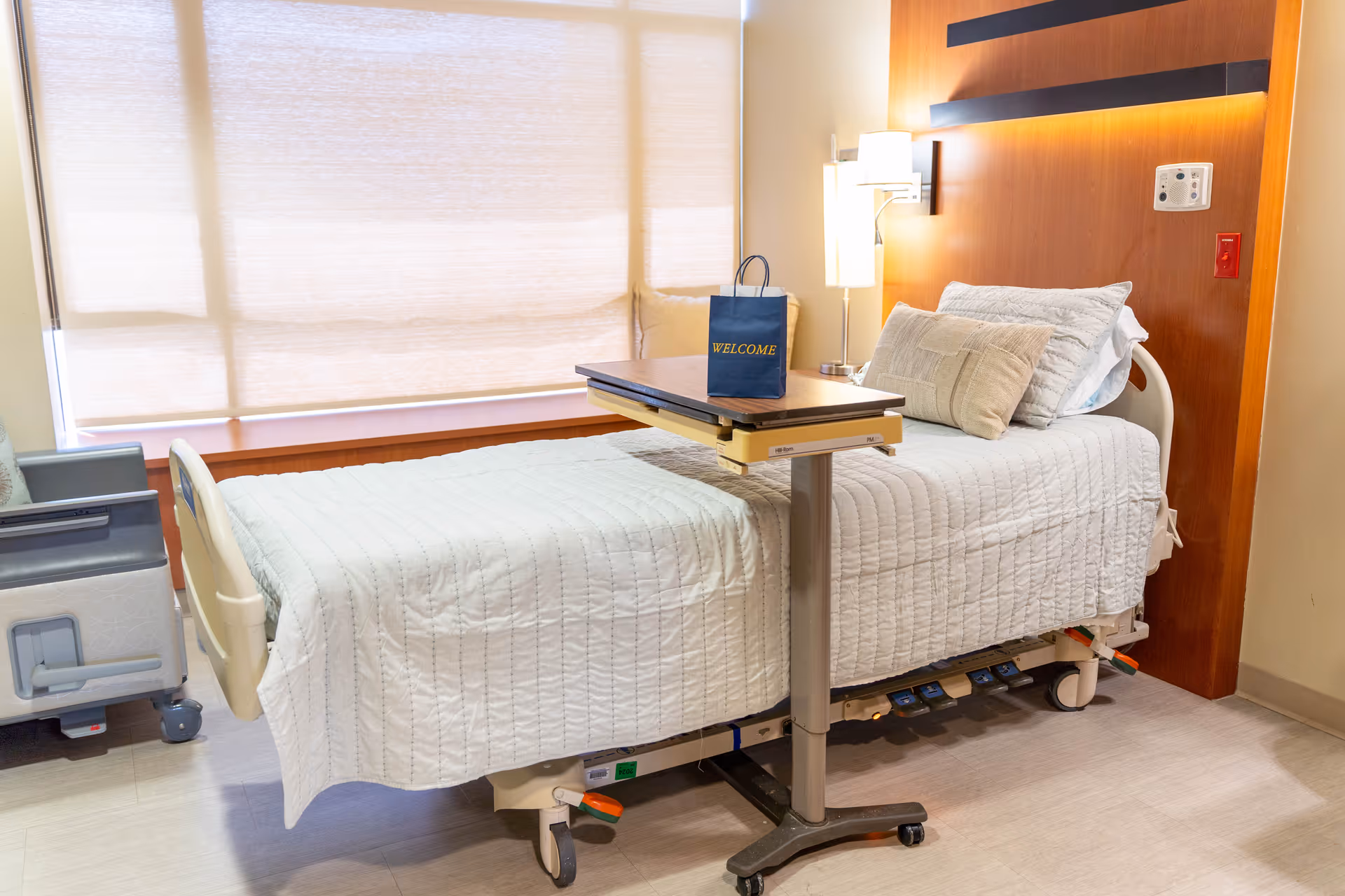A clean and well-lit hospital-style bedroom with a single adjustable bed covered in a white quilt. The bed has several pillows and a small overbed table with a blue bag labeled 'WELCOME' on it. There is a window with closed blinds behind the bed and a bedside lamp attached to a wooden headboard.
