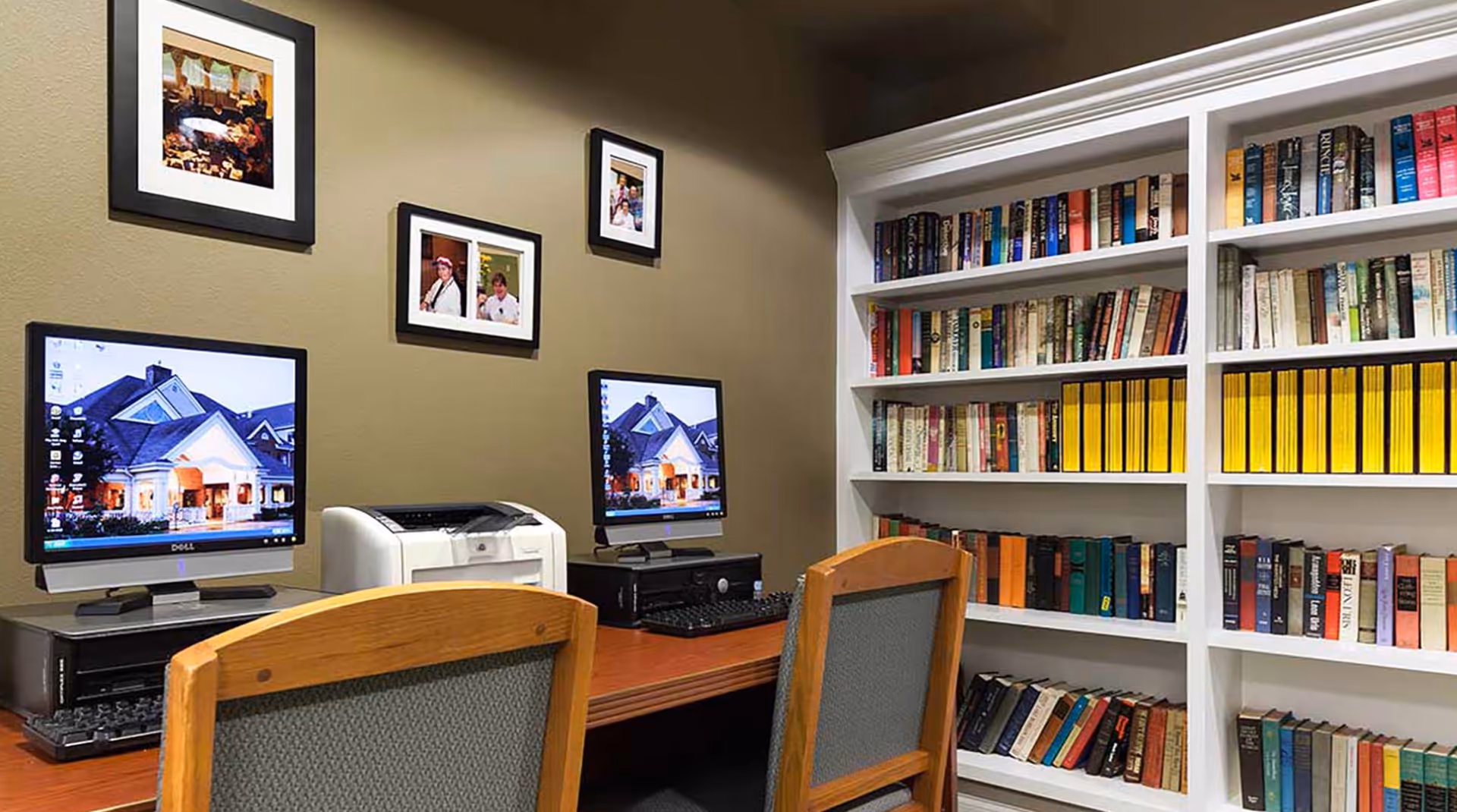 Two computer workstations with monitors and a printer next to a bookshelf filled with books in a small communal library/work area.