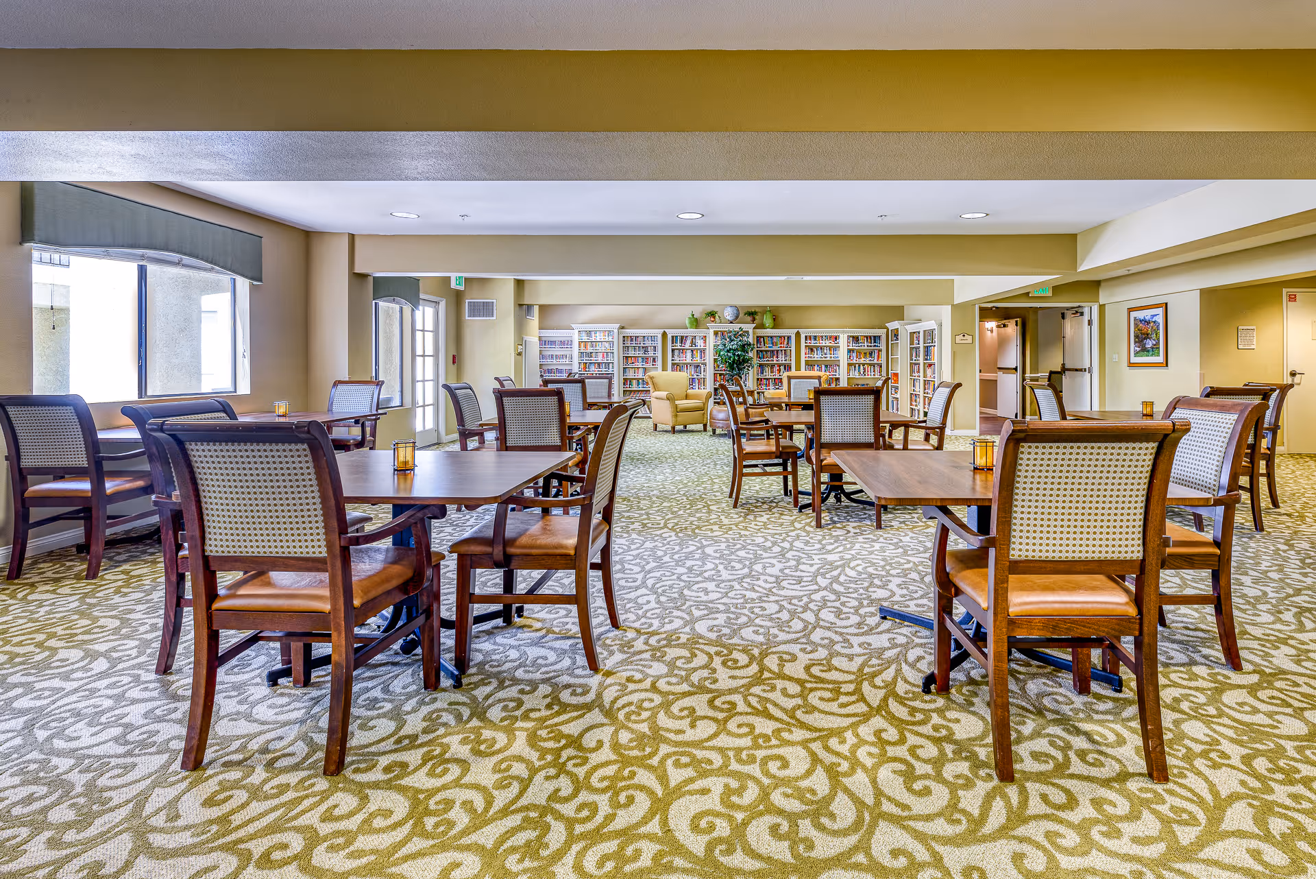 A spacious senior living common area with multiple wooden tables and chairs arranged on a patterned carpet. The room has large windows on the left side allowing natural light to enter. At the far end, there are bookshelves filled with books and a comfortable armchair near a potted plant. The walls are painted in a soft yellow tone, and there are exit signs above the doors.