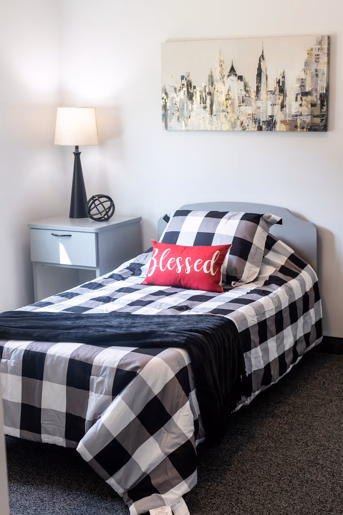 A neatly made single bed with black and white checkered bedding and a red pillow with the word 'Blessed' on it. Next to the bed is a gray nightstand with a black lamp and decorative orb. Above the bed hangs a painting of a city skyline in neutral tones. The room has white walls and dark carpeted flooring.