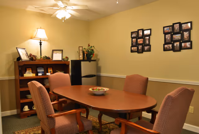 A cozy interior room with a wooden oval table surrounded by four upholstered chairs. On the table is a decorative bowl. Against the wall is a wooden bookshelf with framed pictures, books, and a lamp on top. Next to the bookshelf is a small black refrigerator with a plant on top. The walls are beige with a white chair rail, and two photo collage frames hang on the wall.