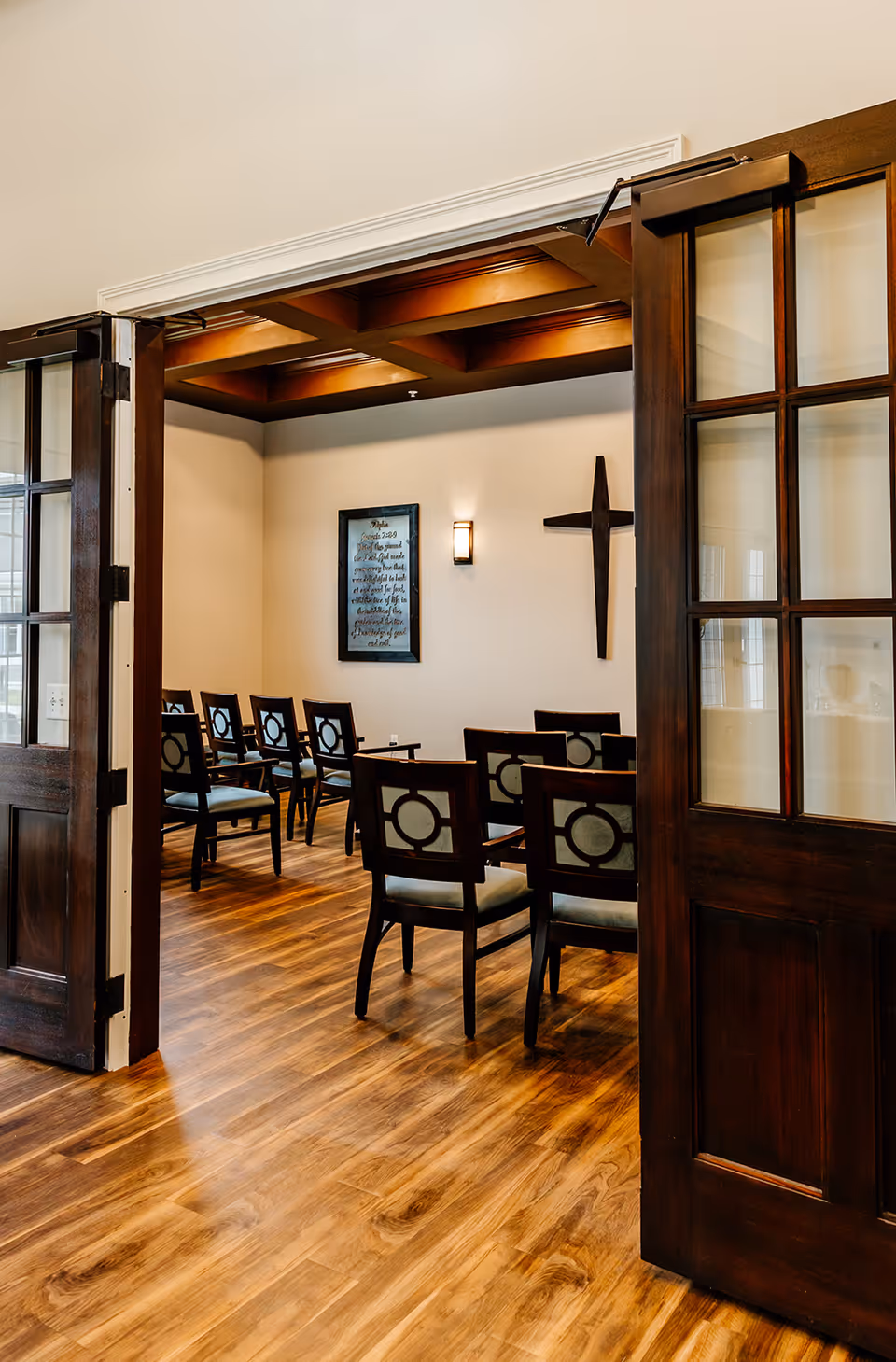 Open double doors reveal a wood-floored interior room with rows of chairs, a wall-mounted cross, and framed artwork.