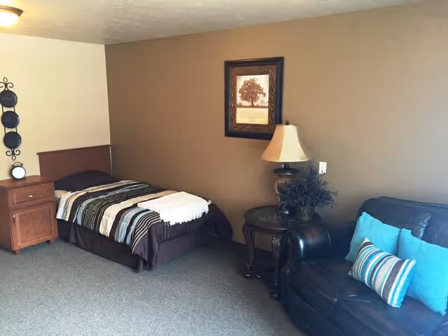 Furnished resident room with a single bed, nightstand, framed art, lamp on a side table, and a leather sofa with pillows against a brown wall.