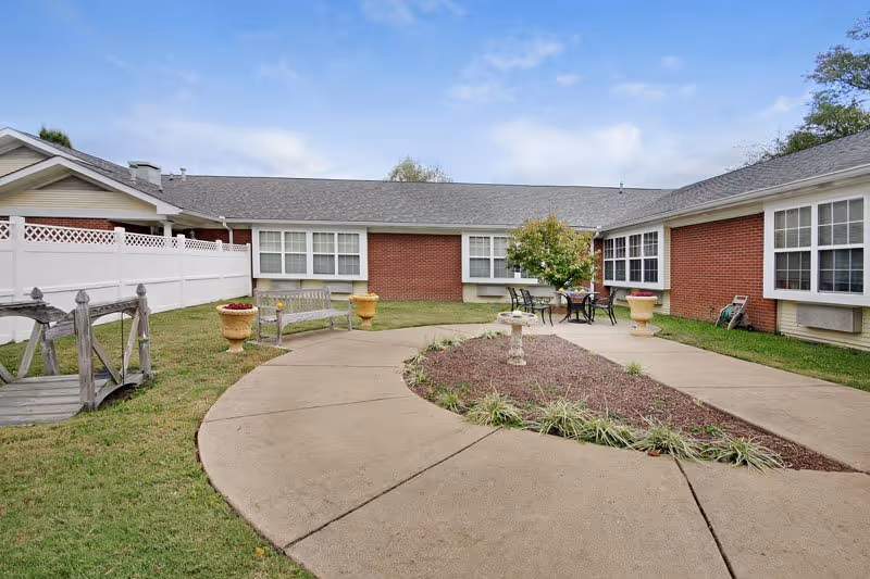 Outdoor courtyard area of a senior living facility with a curved concrete walkway surrounding a mulched garden bed with plants and a birdbath. There are two benches, a small wooden bridge over grass, and a table with four chairs near a small tree. The building has red brick walls and multiple windows, with a white fence on one side and a partly cloudy sky above.