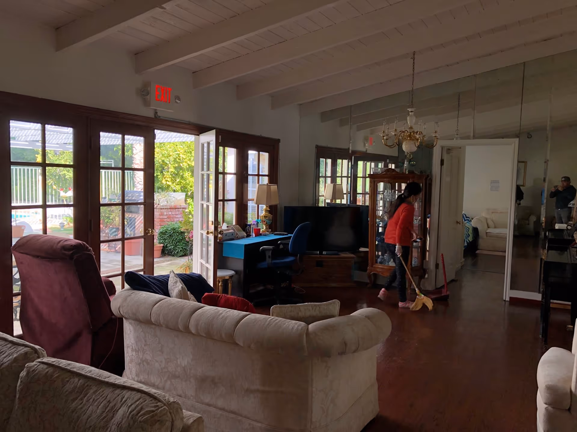 Interior view of a living room with multiple sofas and armchairs. A person wearing a red jacket is sweeping the wooden floor near a glass cabinet and a television. Large glass doors open to an outdoor area with plants and a pool visible. The ceiling has exposed white beams and a chandelier hangs above.