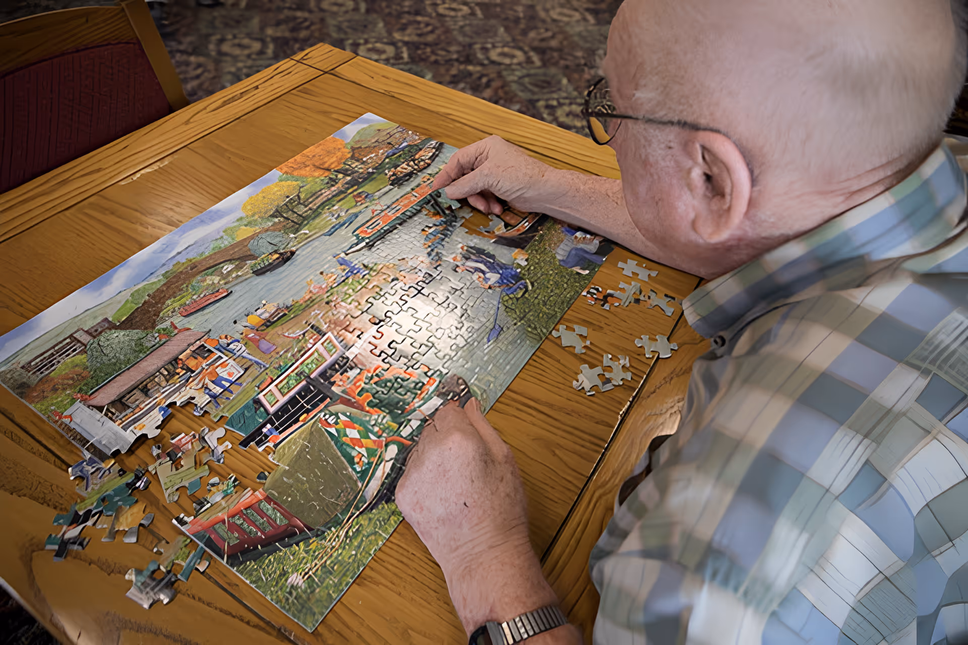 An elderly man wearing glasses and a plaid shirt is sitting at a wooden table working on a colorful jigsaw puzzle depicting a scenic outdoor setting with trees, water, and boats. Several puzzle pieces are scattered on the table nearby.