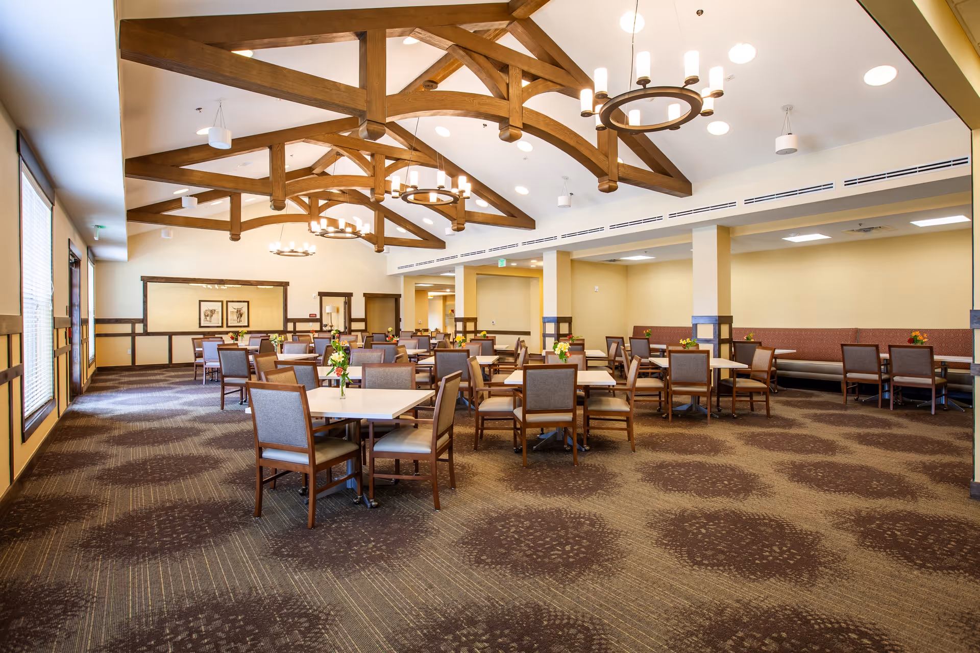 A spacious dining room with multiple square tables and wooden chairs arranged neatly. The room features a high ceiling with exposed wooden beams and circular chandeliers. The walls are painted light beige with wooden trim, and the carpet has a patterned design. Small flower arrangements are placed on each table.