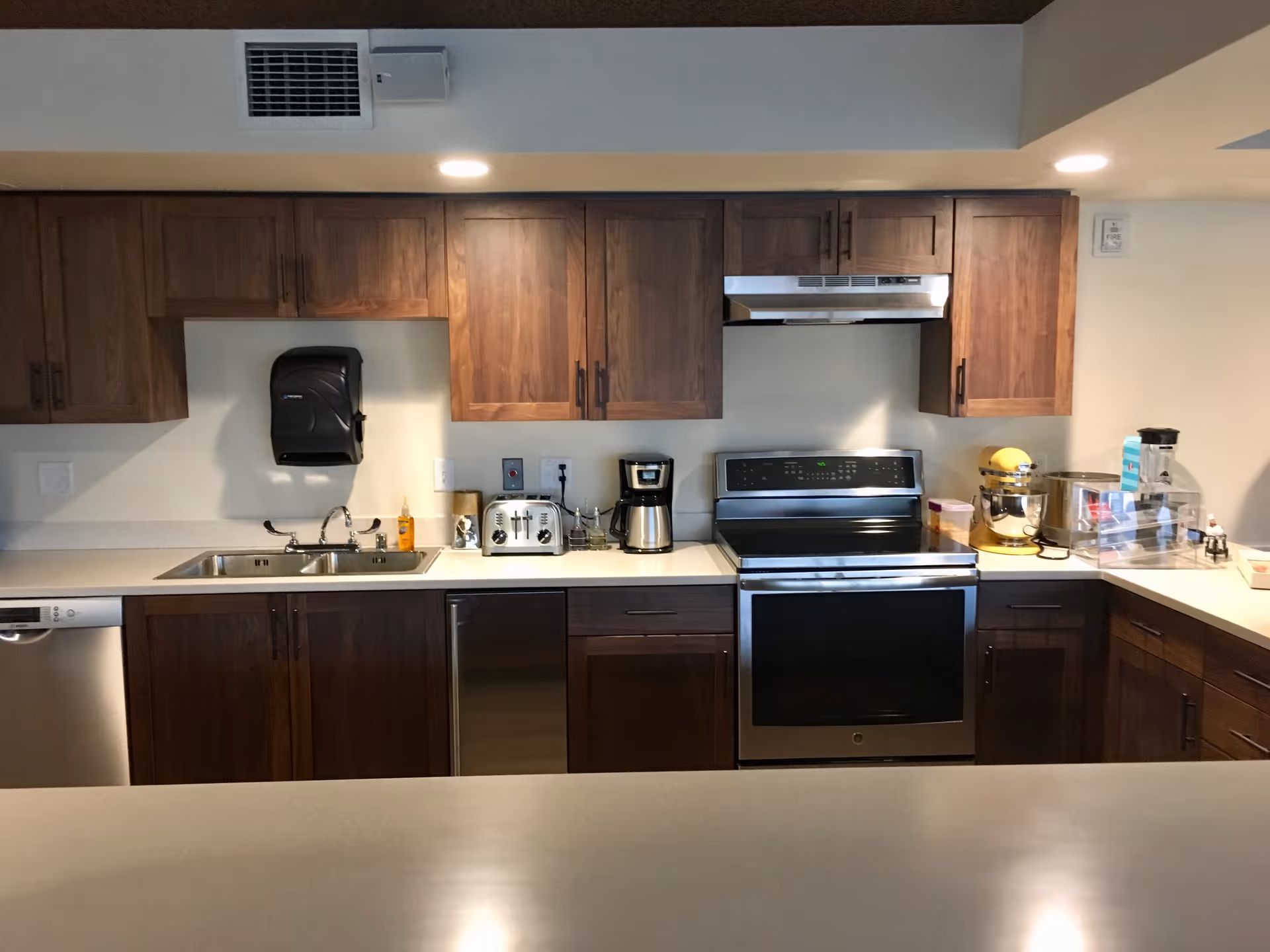 A modern kitchen with wooden cabinets, a stainless steel sink, a dishwasher, a toaster, a coffee maker, an electric stove with an oven, a yellow stand mixer, and various kitchen items on the countertops.