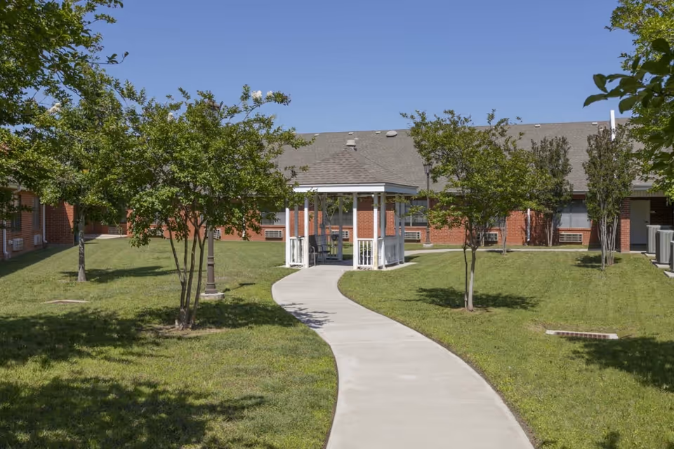 A curved concrete path leads to a white gazebo in a grassy courtyard with trees and a low brick building under a clear blue sky.