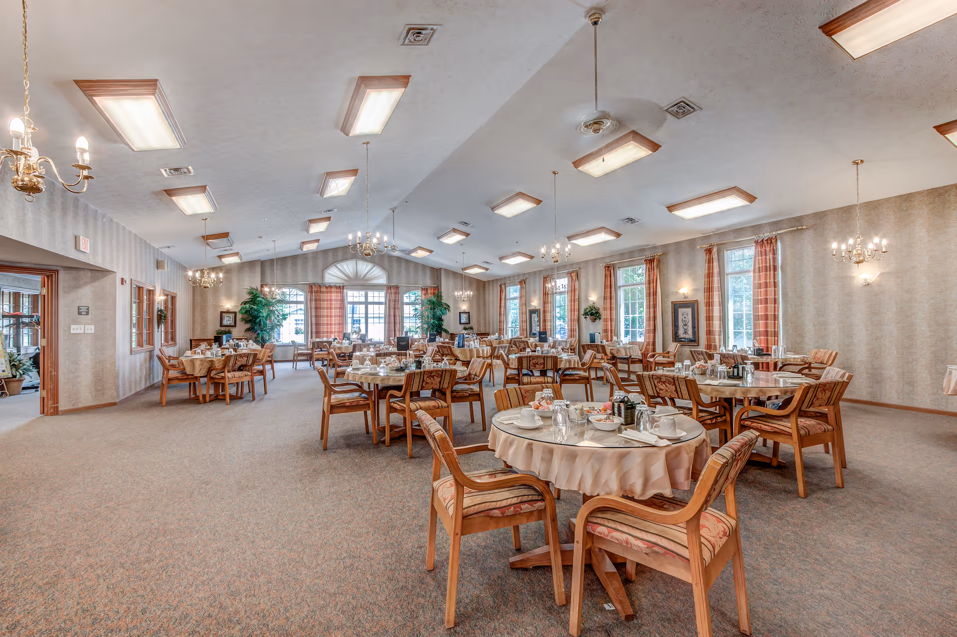 A spacious dining room with multiple round tables covered with tablecloths, set with cups, plates, and utensils. The room has large windows with plaid curtains, chandeliers, and ceiling lights. Wooden chairs with patterned cushions surround the tables. There are plants and framed pictures along the walls.