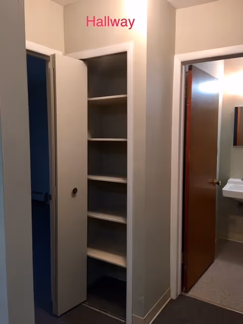Interior hallway with an open linen closet revealing built-in shelves and doors to adjoining rooms, one showing a bathroom sink.