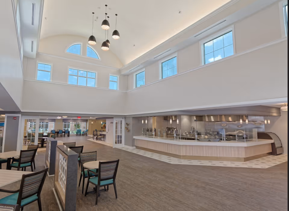Spacious dining area with high ceilings and large windows letting in natural light. Multiple tables and chairs are arranged for seating, with a serving counter and kitchen area visible on the right side. Modern pendant lights hang from the ceiling.