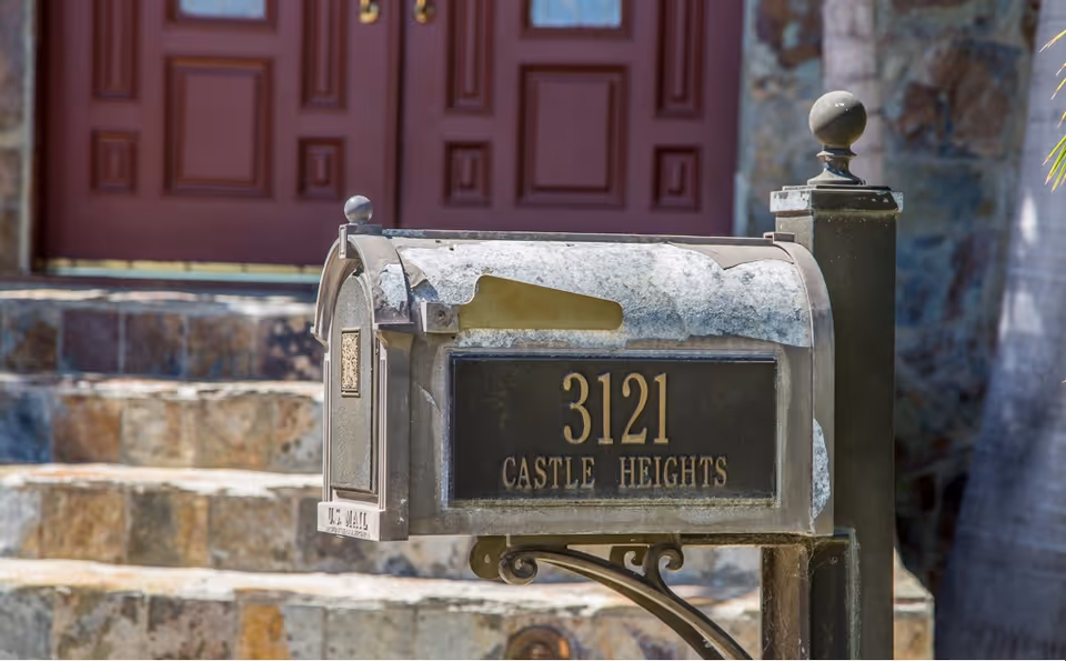 A close-up view of a weathered mailbox with the address number 3121 and the name 'Castle Heights' on it, positioned in front of stone steps leading up to a building with double red doors.