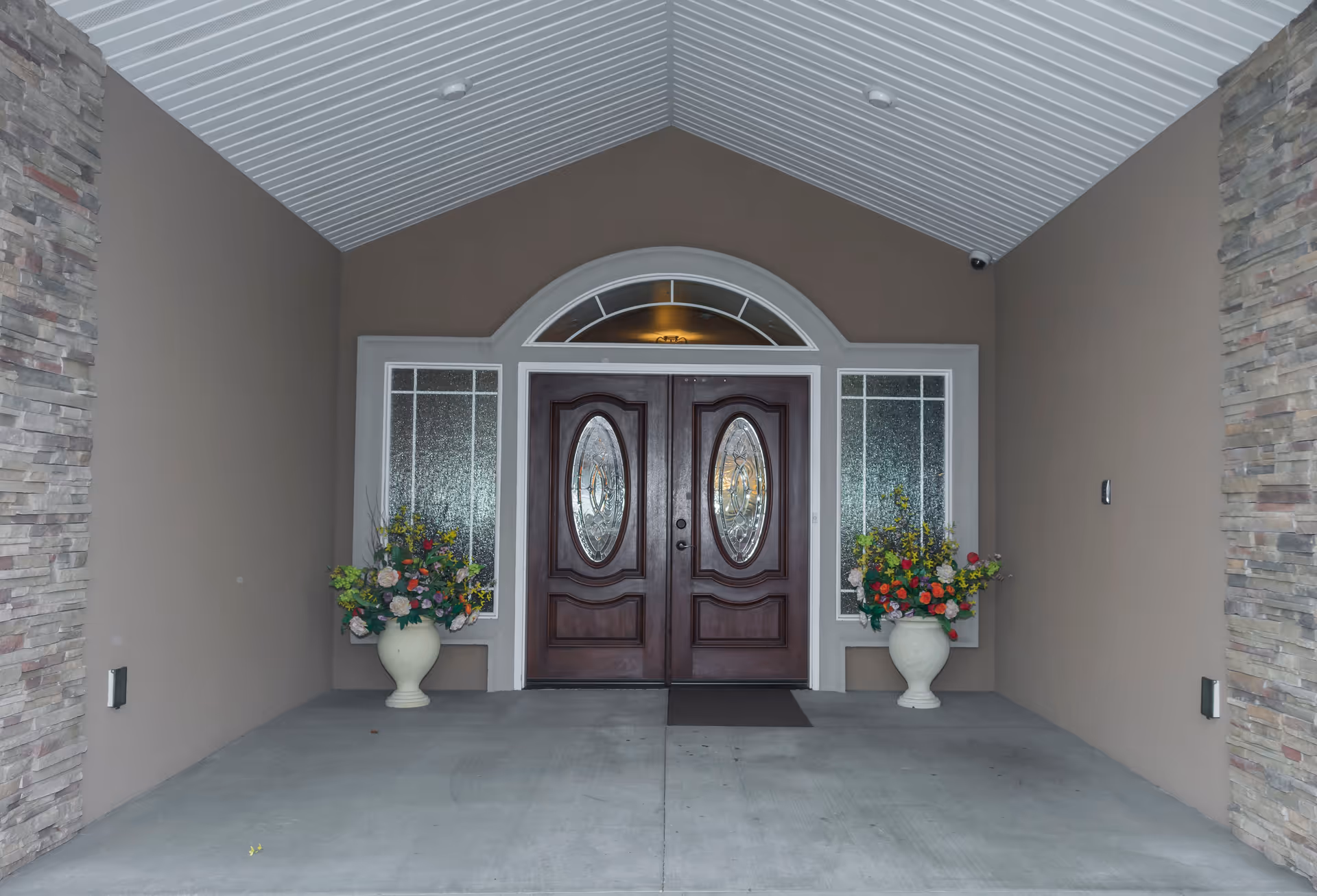 Covered entrance with double wooden doors featuring oval glass panels, flanked by two large flower pots with colorful artificial flowers, stone walls on the sides, and a white paneled ceiling.