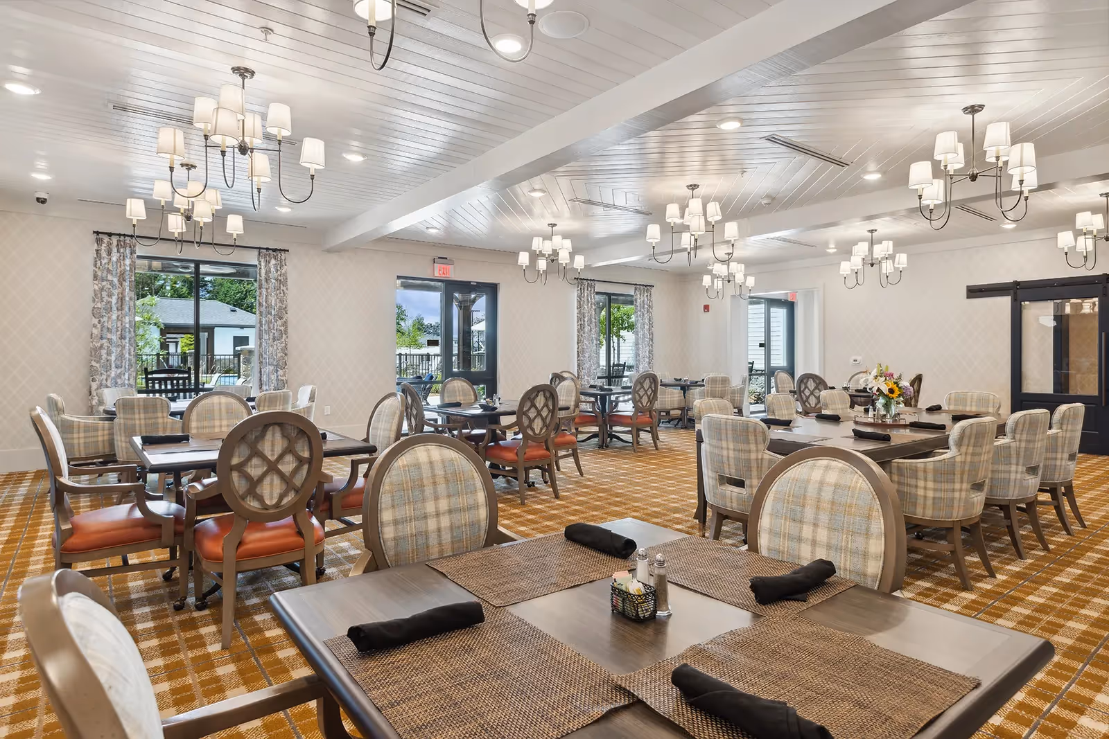 A spacious dining room with multiple tables and chairs arranged neatly. The chairs have patterned upholstery and the tables are set with placemats and black napkins. Large windows with patterned curtains allow natural light to enter, and elegant chandeliers hang from the white paneled ceiling. The room has a warm, inviting atmosphere with a patterned carpet and neutral wall colors.