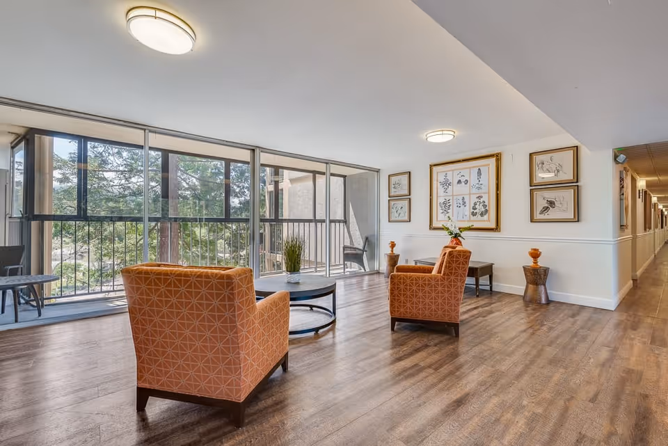 A bright and spacious sitting area in a senior living facility with two orange patterned armchairs facing a round coffee table. Large floor-to-ceiling windows provide a view of trees outside. The walls are decorated with framed artwork, and a long hallway extends to the right.