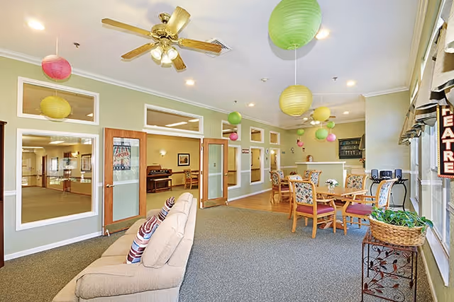 A bright communal lounge with a sofa, round dining tables and chairs, hanging paper lanterns, and large windows.