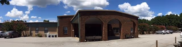 Panoramic exterior view of a single-story brick building with an arched covered entrance, parking lot, and blue sky.
