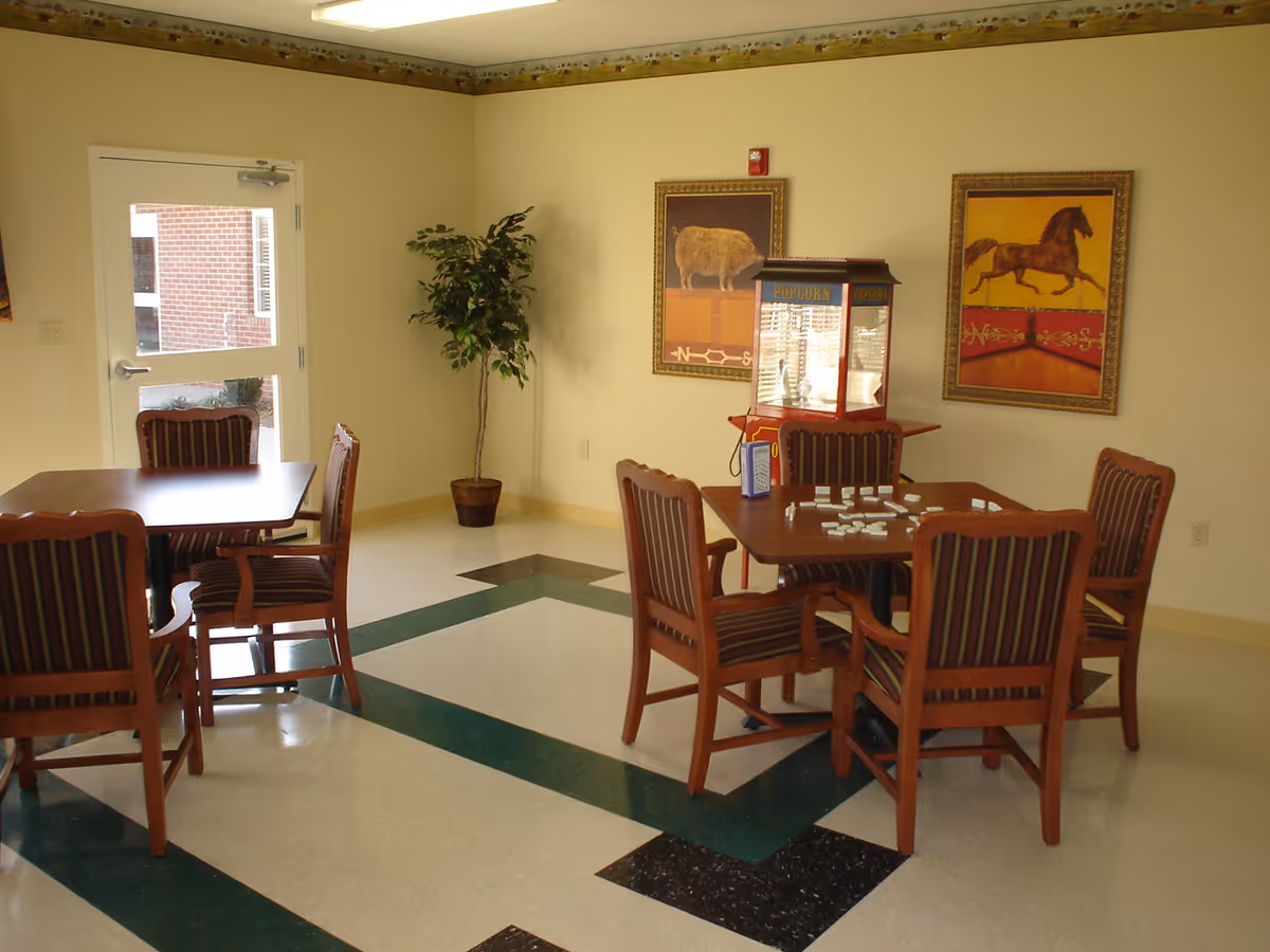 A common area with two wooden tables and striped cushioned chairs. One table has dominoes set up on it. There is a popcorn machine on a small table against the wall. Two framed animal paintings, one of a bison and one of a horse, hang on the wall. A potted plant is in the corner near a door with a window showing a brick exterior.