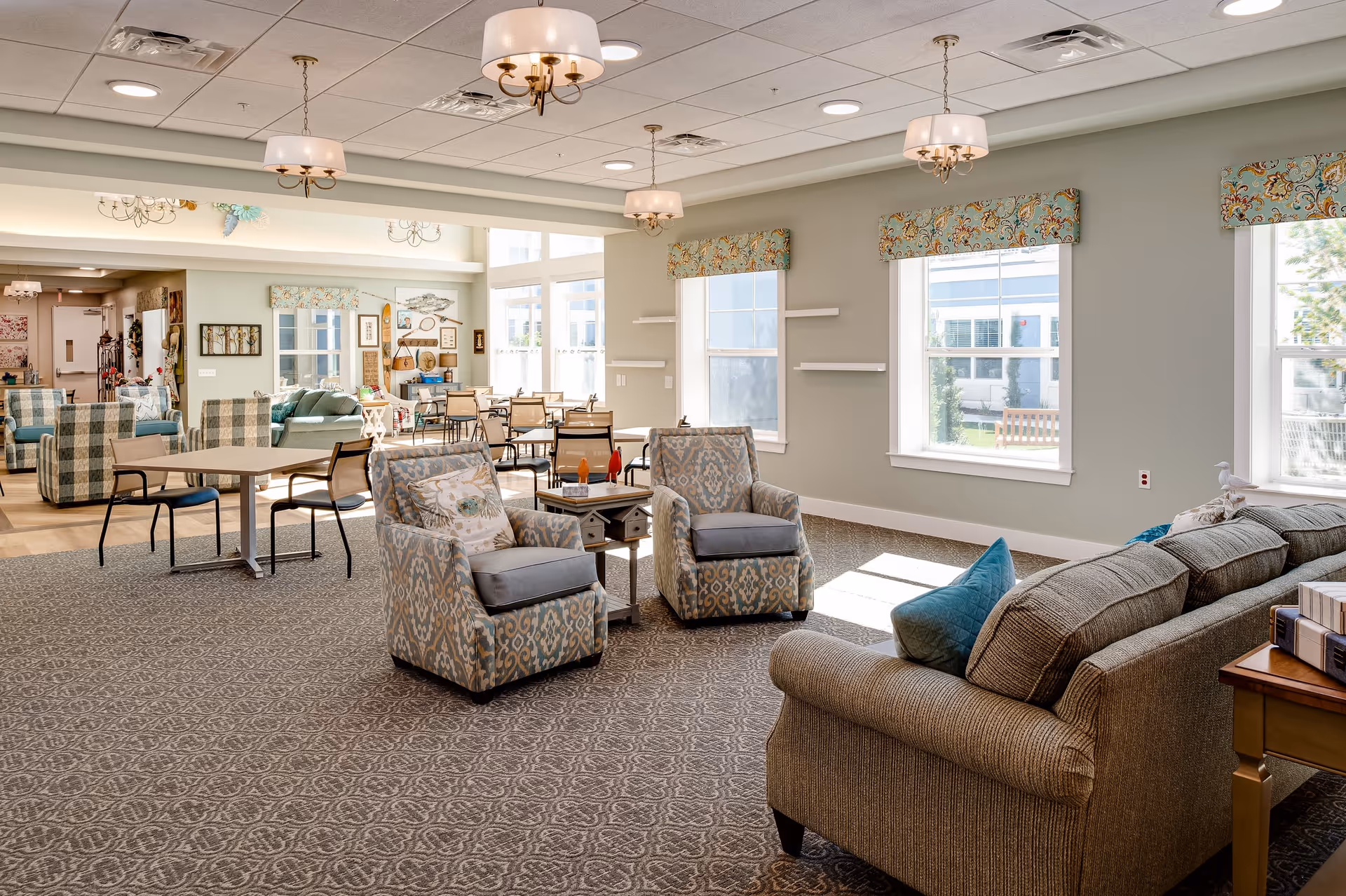 A bright and spacious common area in a senior living facility featuring patterned armchairs, a sofa with blue cushions, several tables with chairs, large windows with decorative valances, and elegant ceiling light fixtures. The room has a carpeted floor and light-colored walls with some wall decorations and shelves.